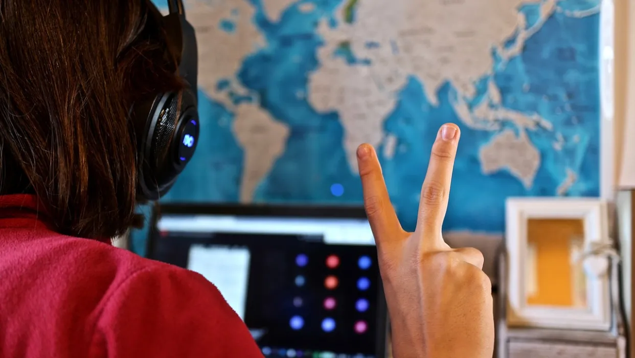 Student with headphones on looking at a laptop screen holding fingers up in peace symbol